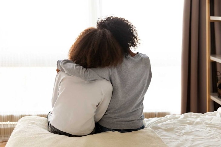 Rear back view black mother and daughter embrace sitting on bed at home, older sister consoling younger teen, girl suffers from unrequited love share secrets trustworthy person relative people concept