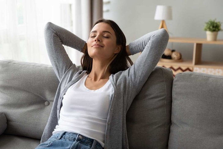 Relaxed serene pretty young woman feel fatigue lounge on comfortable sofa hands behind head rest at home, happy calm lady dream enjoy wellbeing breathing fresh air in cozy home modern living room