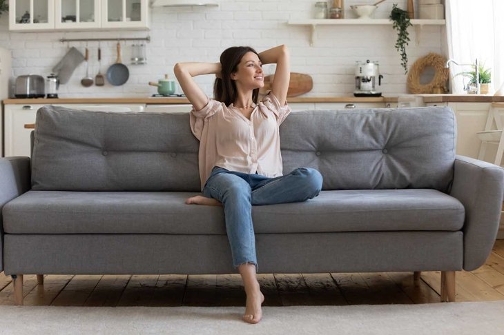 In cozy living room happy woman put hands behind head sitting leaned on couch 30s european female enjoy lazy weekend or vacation, housewife relaxing feels satisfied accomplish chores housework concept