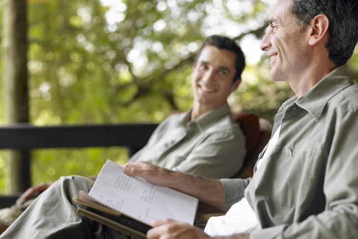 Side view of a happy man with book sitting on terrace with blurred male friend in background